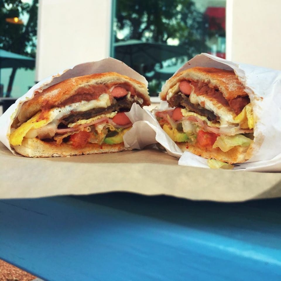 A Mexican torta, cut in half, revealing layers of meat, hot dogs, egg, and vegetables.